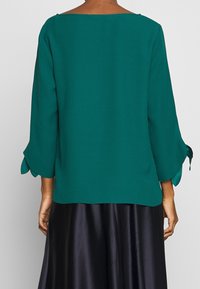 Blouse bleu sarcelle à manches trois-quarts et encolure dégagée, avec des liens ruban aux extrémités des manches. Tissu lisse et léger.