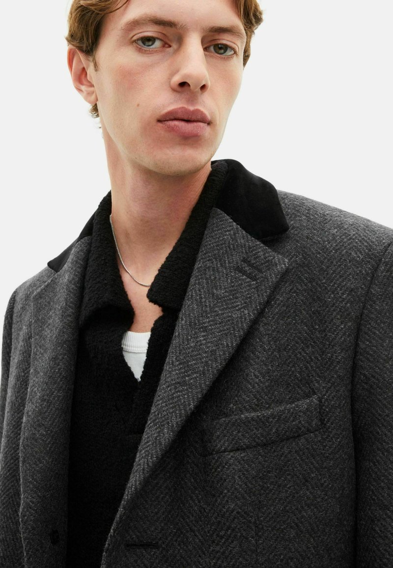 Grey herringbone coat with black velvet collar, layered over a black textured sweater with a white crew neck. Simple silver chain visible.