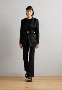 Svart satengblazer med belte, kombinert med tettsittende svarte vide jeans og plattform sandaler. Glatte teksturer og skreddersydd design.