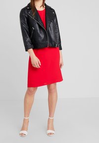 Chaqueta de biker de cuero negro con cremalleras plateadas sobre un vestido rojo hasta la rodilla; la modelo lleva sandalias de tacón blancas. Texturas suaves, diseño casual.