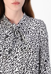 Schwarz-weiße Bluse mit geometrischem Muster und Halsbinde, die verschiedene dreieckige und rechteckige Formen aus einem glatten Stoff zeigt.