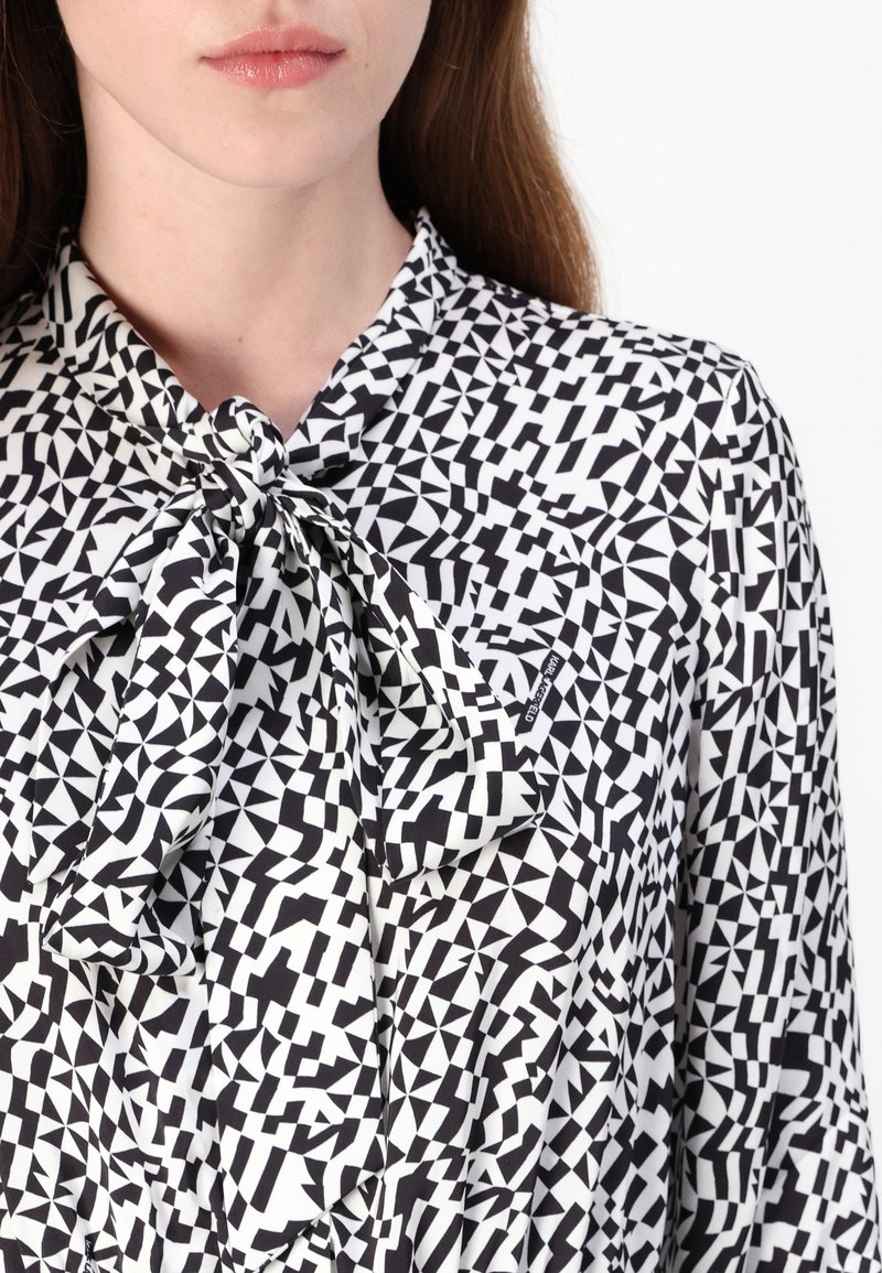 Schwarz-weiße Bluse mit geometrischem Muster und Halsbinde, die verschiedene dreieckige und rechteckige Formen aus einem glatten Stoff zeigt.