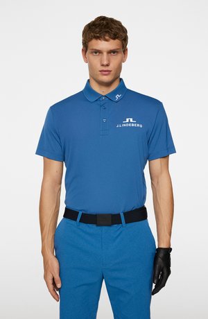 Blue polo shirt with a collar, featuring a logo on the chest. Paired with light blue textured pants and a black belt. Black glove on left hand.