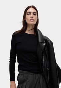 Schwarzes Langarmshirt mit taillierter Passform, getragen unter einer schwarzen Lederjacke. Das Model hat glattes, dunkles Haar und trägt graue Hosen.