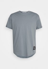 Camiseta gris claro de manga corta con cuello redondo y pequeño parche negro con logo cerca del dobladillo sobre un fondo blanco.