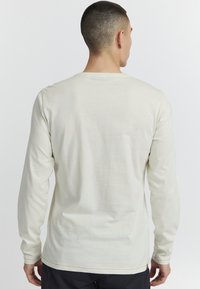 Camisa de manga larga en color crema claro, hecha de una tela suave. Tiene un cuello redondo, acentos cosidos y un ajuste relajado.
