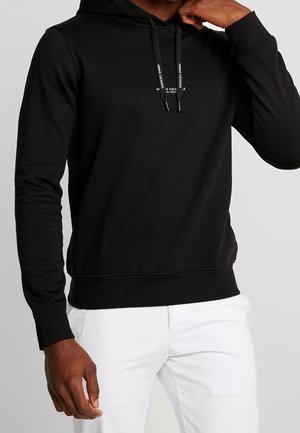 Man wearing black hoodie with Armani Exchange logo on drawstrings and white pants, hand near neck, against plain background.
