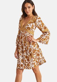 Robe courte avec de longues manches cloche, présentant un décolleté en V profond. Motif floral en moutarde et marron sur fond blanc.