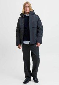 Gewatteerde zwarte jas met capuchon, voorzien van twee voorkantzakken en een ritssluiting, gedragen over een navy trui en gestreept overhemd, gecombineerd met grijze broek.