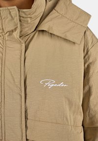 Beige gesteppte Jacke mit hohem Kragen, die eine dezente Struktur aufweist und ein weißes gesticktes Logo auf der Brust trägt.