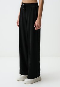 Zwarte, wijde broek met een elastische tailleband, trekkoorddetail en zachte textuur; gecombineerd met witte sneakers.