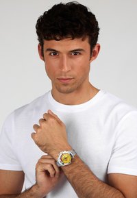 Orologio da polso argento con cassa rotonda, quadrante giallo, dettagli neri e cinturino in acciaio inossidabile, indossato su un polso nudo.