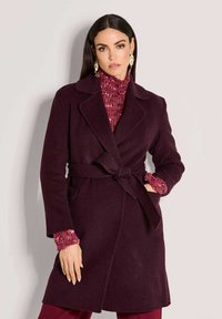 Vrouw in een donkerpaarse ceintuurjas over een rode gebreide trui en rode broek, poserend met één hand in de zak tegen een effen achtergrond.