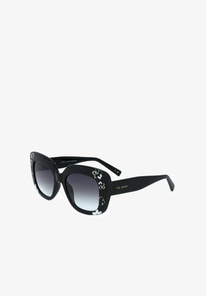 Occhiali da sole neri oversize a forma di occhi di gatto con accenti floreali bianchi e lenti scure sfumate su sfondo bianco.