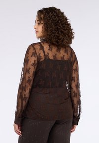 Blusa marrone trasparente con motivo floreale in pizzo, maniche lunghe e orlo arrotondato. Presenta una scollatura quadrata e polsini con bottoni.