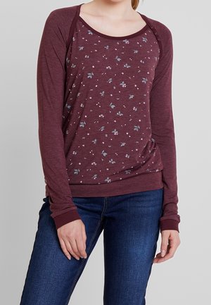 Kobieta w długim rękawie bordowej bluzki z małymi kwiatowymi wzorami i ciemnoniebieskich jeansach, stojąca z rękami luźno opuszczonymi wzdłuż ciała.