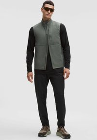 Muž v tmavých slnečných okuliaroch, šedý veste na zips, čiernej dlhý rukáv, čierne nohavice a béžovo-hnedé tenisky stojaci na jednofarebnom pozadí.