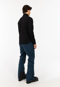 Zwarte lange-mouwen top met een hoge kraag, gecombineerd met teal sneeuwbroek met ritszakken en donkere stofaccenten; zwarte sneeuwschoenen zichtbaar.