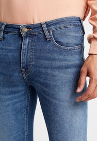 Blå denimjeans med en åtsittande design, som har dragkedja och metallknappstängning. Lätt blekning och sömnadsdetaljer på fickorna.