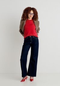 Νεαρή γυναίκα με σγουρά μαλλιά, φοράει γυαλιά, καφέ σακάκι, κόκκινο τοπ, σκουρόχρωμο τζιν με φαρδιά πόδια και κόκκινα ψηλοτάκουνα παπούτσια, στέκεται μπροστά σε απλό φόντο.