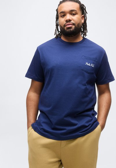 Άνδρας με τα χέρια στις τσέπες, ντυμένος με μπλε σκούρο μπλουζάκι Polo RL και μπεζ παντελόνι, στέκεται μπροστά σε ανοιχτό γκρι φόντο.