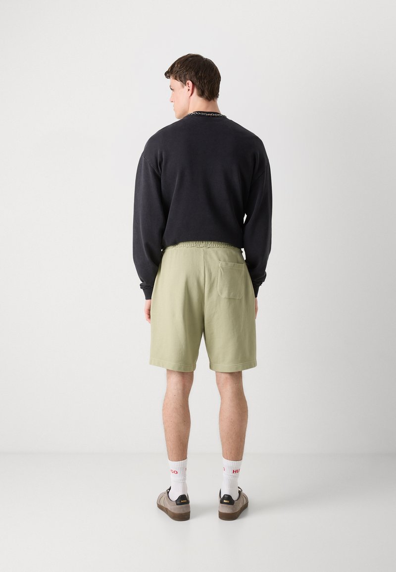 Schwarzes Sweatshirt mit gerippten Bündchen, grüne Shorts mit elastischem Bund und Gesäßtasche, weiße Socken mit roter Schrift, braune Turnschuhe.