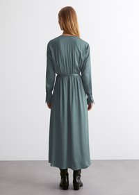 Lang, teal satengkjole med innsnevret midje, lange ermer og knappede mansjetter. Glatt tekstur og flytende silhuett, som når midt på leggen.