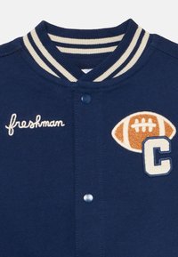 Marineblå varsity-jakke med kremfargede striper på kragen, brodert "freshman," og et fotballmerke med "C"-detalj. Jeg har blå trykknapper.