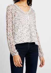 Lehká, průsvitná blůza s výstřihem do V, dlouhými rukávy a nařasenými manžetami. Má květinový vzor v pastelových barvách na bílém pozadí.