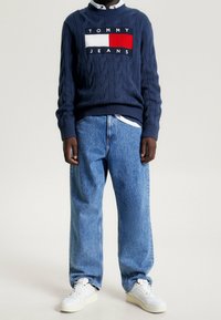 Marinblå kabelstickad tröja med vit, röd och svart logotyp; matchad med ljusblå baggy jeans och vita sneakers.