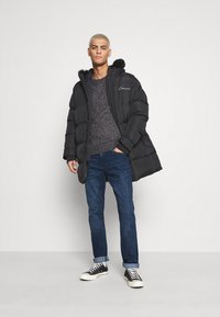 Zwarte pufferjack met een bont gevoerde capuchon, gedragen over een gestructureerde grijze trui, gecombineerd met donkere jeans en zwarte sneakers met witte zolen.