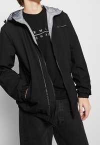 Svart vindjacka med gråfärgad foder på huven, elastiska ärmmuddar och en logotypdetalj. Har dragkedjestängning och en lätt, slät textur.