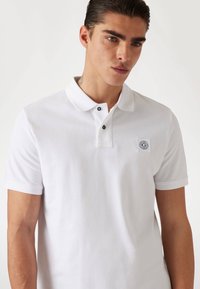 Weißes Poloshirt aus weichem Stoff, mit klassischem Kragen, einer Knopfleiste mit drei schwarzen Knöpfen und einem kleinen Logo-Patch auf der Brust.