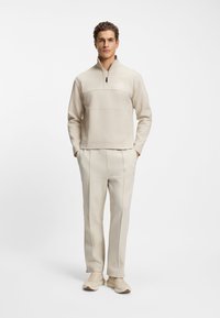 Beżowa bluza z suwakiem do ćwiartki z teksturowanym górnym panelem, zestawiona z dopasowanymi, zwężanymi spodniami. Oba elementy cechuje minimalistyczny design i subtelne oznakowanie.