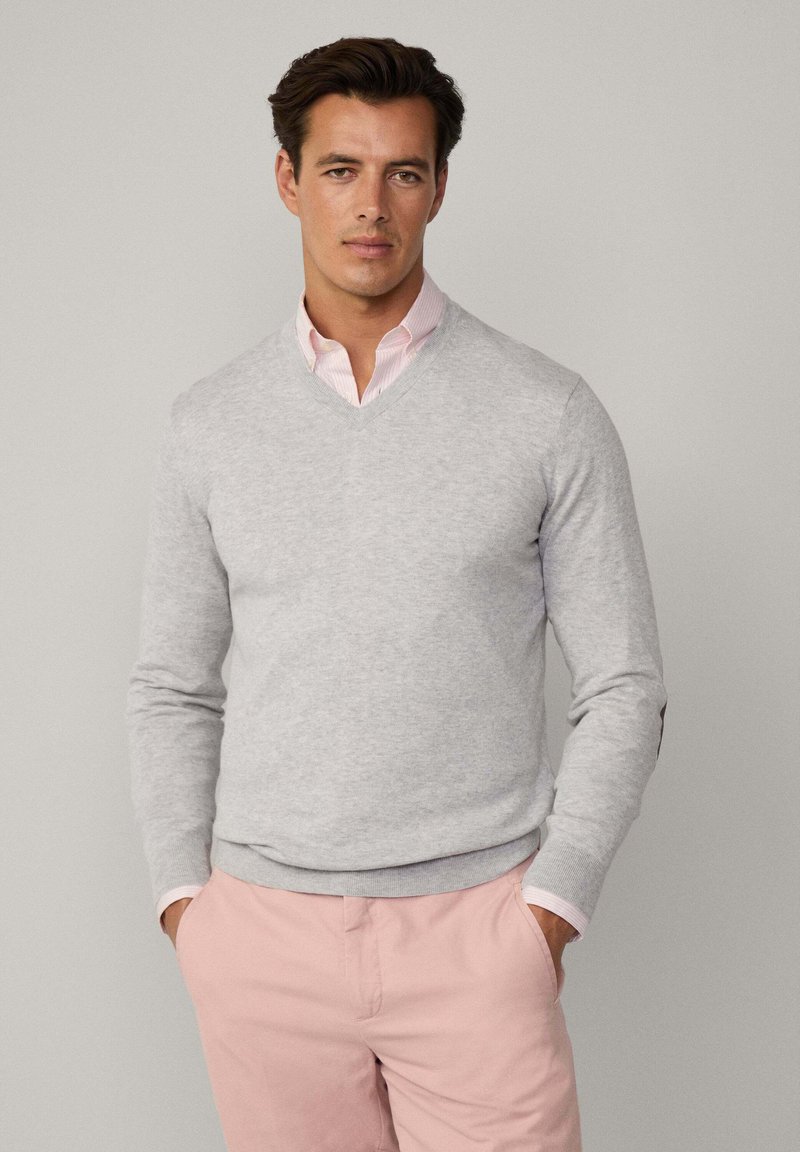 Grauer V-Ausschnitt-Pullover aus weichem Material, mit langen Ärmeln und gerippten Bündchen, getragen über einem hellrosa, gestreiften Hemd mit Kragen.