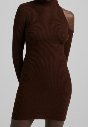 Robe en maille brun foncé, près du corps, côtelée, avec un col montant et une découpe à une épaule, portée par une personne sur un fond gris uni.