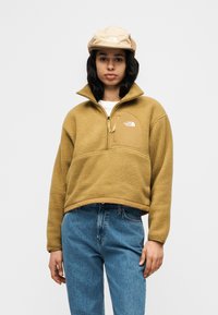 Φλις πουλόβερ σε ταμπά χρώμα με φερμουάρ στο ένα τέταρτο, ψηλό γιακά και μπροστινή τσέπη. Συνδυάζεται με ανοιχτό γαλάζιο τζιν παντελόνι. Το καπέλο είναι μπεζ με λογότυπο.