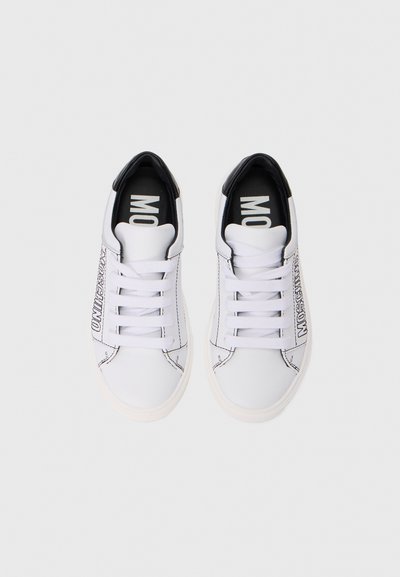 Vita sneakers med svarta detaljer, platta snören och en kontrasterande logotyp på sidan. Tillverkade av slätt material, med rundad tå.
