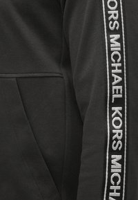 Czarna bluza z gładką fakturą, z wyraźnym białym i srebrnym logo "MICHAEL KORS" biegnącym wzdłuż bocznego szwu.