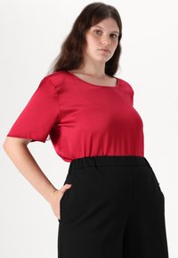 Punane satiinist lühikeste käistega pluus ümara kaelusega, koos mustade kõrge vöökohtadega pükstega. Kanga pind näib sile ja kerge läikega.