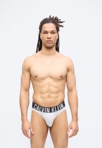 Άνδρας χωρίς μπλούζα με κότσους dreadlocks στέκεται μπροστά, φοράει λευκά εσώρουχα Calvin Klein με μαύρη ελαστική ζώνη.