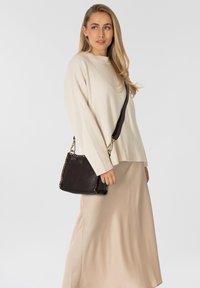 Donna che indossa un maglione beige e una gonna, tiene in mano una borsa nera con borchie dorate, mentre guarda leggermente di lato su uno sfondo bianco.