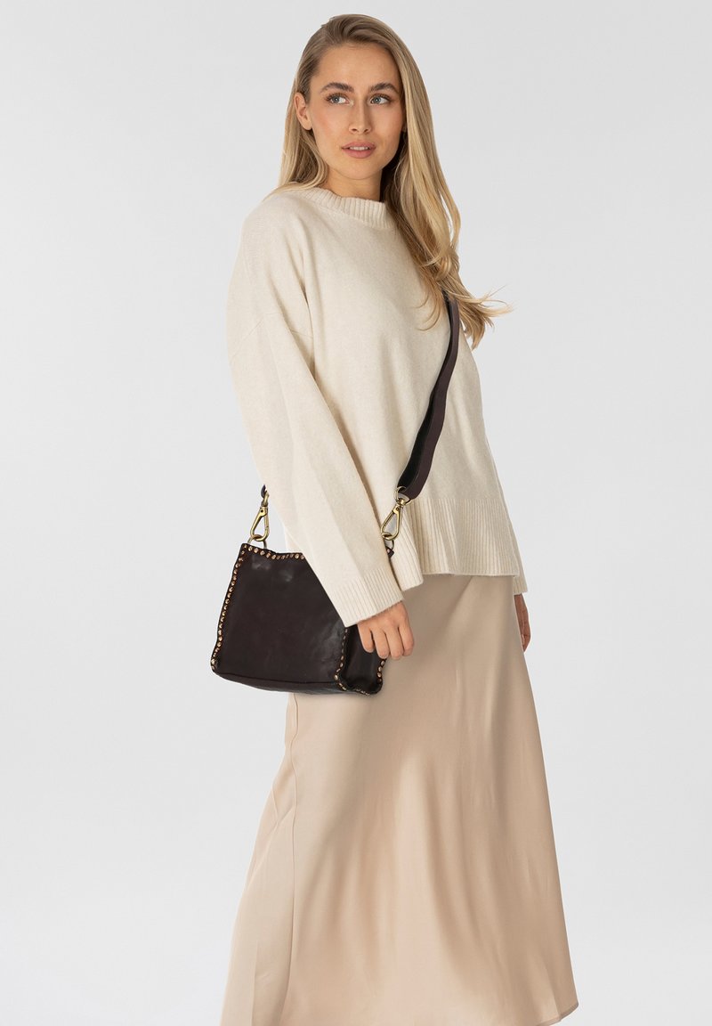 Donna che indossa un maglione beige e una gonna, tiene in mano una borsa nera con borchie dorate, mentre guarda leggermente di lato su uno sfondo bianco.