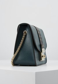 Borsa in pelle color verde scuro con tracolla in catena dorata, accentata da pelle nera chiodata e chiusura a lucchetto dorato, poggiata su una superficie bianca.