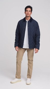 Un giovane indossa una giacca trapuntata blu navy, una camicia bianca, pantaloni beige e sneakers bianche davanti a uno sfondo semplice.