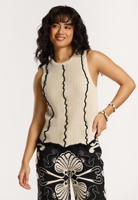 Adīta bežīga bez piedurknēm blūze ar melnu viļņotu apdari, teksturēta virsma un brīvs griezums; kombinēta ar rakstainām melnām biksēm ar baltām zīmēm.