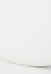 Bela usnjena dodatka s tiskanim logotipom "JOOP! JEANS". Teksturirana površina z gladkim zaključkom. Krožna oblika s šivom.