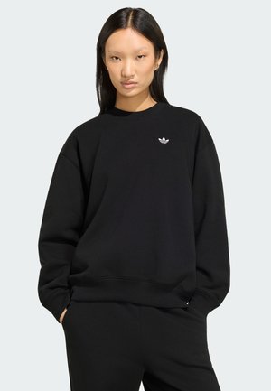 Młoda kobieta z długimi czarnymi włosami, ubrana w czarną bluzę Adidas i czarne spodnie, stoi z rękami w kieszeniach.