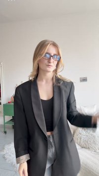 Czarna marynarka z satynowymi klapami, noszona na czarnym krótkim topie. Szare spodnie z teksturowanej tkaniny. Okrągłe okulary z niebieskim odcieniem, średniej długości blond włosy.