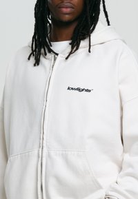 Béžová mikina s kapucňou na zips vyrobená z mäkkej tkaniny; obsahuje predné vrecko a vyšitý logotyp "lowlights" na ľavej strane hrudníka.